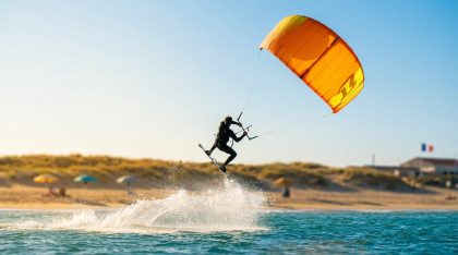 Kitesurfeur effectuant un saut avec son aile orange au-dessus de la mer près d’une plage ensoleillée.