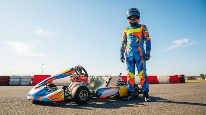 Jeune pilote en combinaison colorée posant debout à côté de son kart sur un circuit en plein soleil.